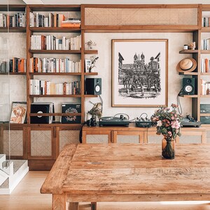 May include: A wooden table with a brown wood top and a light wood base. There is a vase with flowers on the table. The table is in front of a bookshelf with books and a record player. There is a framed black and white drawing of a city scene on the wall behind the table.
