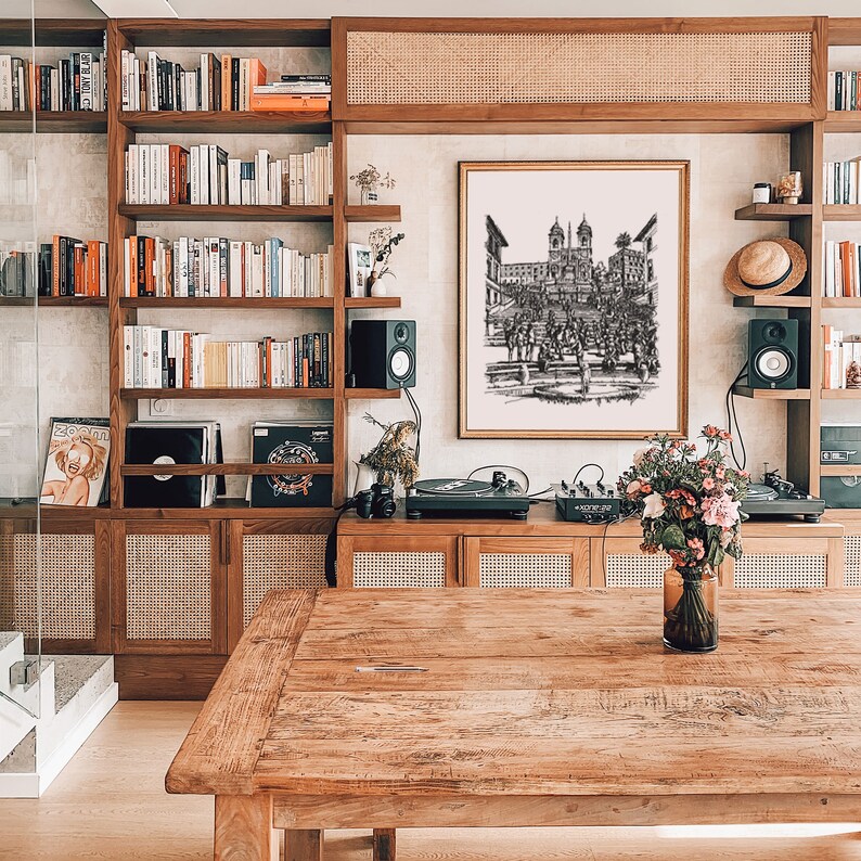 May include: A wooden table with a brown wood top and a light wood base. There is a vase with flowers on the table. The table is in front of a bookshelf with books and a record player. There is a framed black and white drawing of a city scene on the wall behind the table.