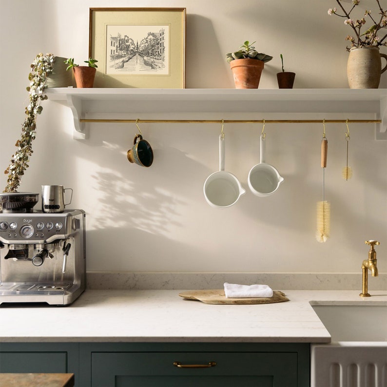 Puede incluir: Una cocina blanca con una estanter&iacute;a blanca con ganchos de lat&oacute;n que sostienen dos cacerolas de esmalte blanco, un cepillo de madera y un cepillo de limpieza de lat&oacute;n. Una m&aacute;quina de espresso Sage se encuentra en la encimera debajo de la estanter&iacute;a.