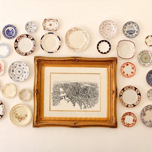 May include: A white wall with a framed black and white print of a city skyline. The print is surrounded by a variety of decorative plates in various colors and patterns.