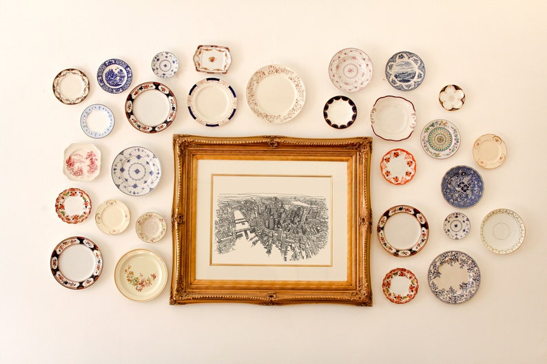 May include: A white wall with a framed black and white print of a city skyline. The print is surrounded by a variety of decorative plates in various colors and patterns.