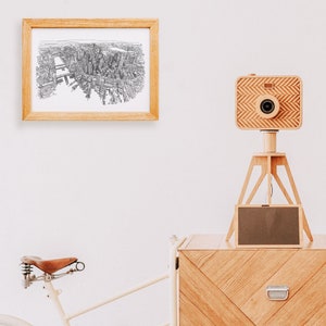 May include: A wooden camera on a tripod with a black and white drawing of a city in a frame on the wall behind it. A wooden chest with a chevron pattern is in the foreground.