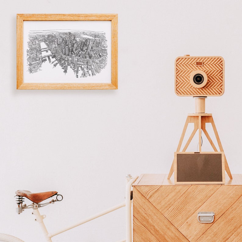 May include: A wooden camera on a tripod with a black and white drawing of a city in a frame on the wall behind it. A wooden chest with a chevron pattern is in the foreground.