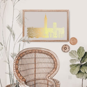 May include: A gold foil print of a city skyline with a tall building in the center, framed in a light wood frame and hanging on a white wall. A wicker chair and a potted plant are in the foreground.