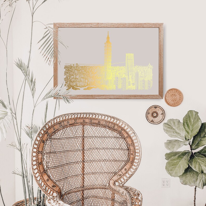 May include: A gold foil print of a city skyline with a tall building in the center, framed in a light wood frame and hanging on a white wall. A wicker chair and a potted plant are in the foreground.