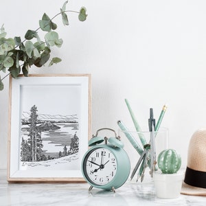 May include: A light green alarm clock with black hands and numbers sits on a white marble surface next to a framed black and white drawing of a lake and mountains. A glass jar filled with pens and pencils is next to the clock, and a small potted cactus is in front of the jar. A straw hat with a black band sits on the right side of the image.