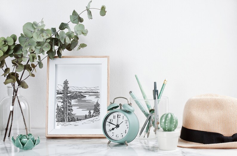 May include: A light green alarm clock with black hands and numbers sits on a white marble surface next to a framed black and white drawing of a lake and mountains. A glass jar filled with pens and pencils is next to the clock, and a small potted cactus is in front of the jar. A straw hat with a black band sits on the right side of the image.