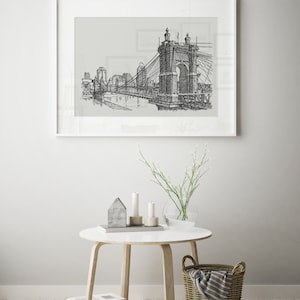 May include: Black and white line drawing of a bridge over a river with buildings in the background. The drawing is framed in a white frame and hanging on a white wall.