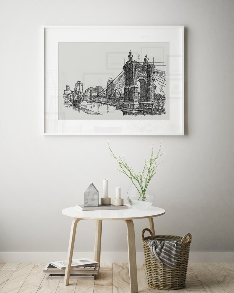 May include: Black and white line drawing of a bridge over a river with buildings in the background. The drawing is framed in a white frame and hanging on a white wall.