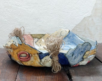 May include: A ceramic bowl with a colorful, abstract design. The bowl is decorated with blue, red, yellow, and white paint. It has a rough, textured surface and appears to be handmade.