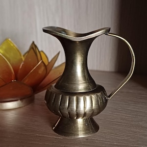 Op de afbeelding: Een kleine, antieke messing kan met een gebogen handvat en een uitlopende tuit. De kan staat op een houten plank, met een decoratieve lotusbloem en boeken op de achtergrond. De kan is ongeveer 10 cm hoog.