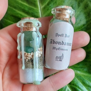 Peut inclure: Deux petits pots en verre avec des bouchons en liège, l'un rempli de cristaux verts, d'herbes et de graines, et l'autre avec une étiquette qui indique "Spell Jar Abundance MyGrimoire".