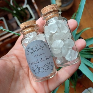 Puede incluir: Dos pequeñas botellas de vidrio llenas de cristales de cuarzo transparentes. Las botellas tienen tapones de corcho y etiquetas que dicen "My Éimeon Cristal de Roche".