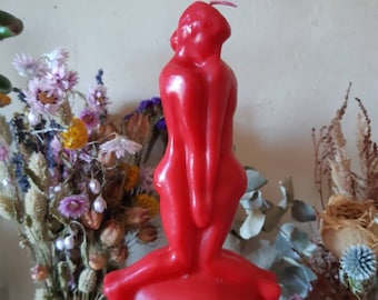 Op de afbeelding: Een rode kaars in de vorm van twee figuren die elkaar omhelzen. De kaars is gemaakt van glad, massief was en heeft een eenvoudig, elegant ontwerp.