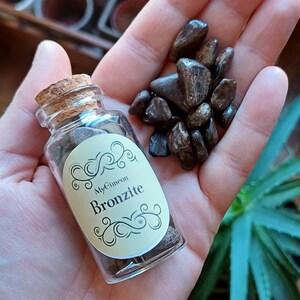 Può includere: Un piccolo flacone di vetro con un tappo di sughero riempito di pietre marroni e nere. Il flacone ha un'etichetta con la scritta "MyCimeon Bronzite".