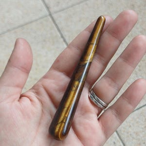 Op de afbeelding: Een gepolijste, cilindrische tijgeroog edelsteen pijp. De pijp heeft een rijke bruine kleur met gouden en zwarte strepen. De edelsteen wordt in een open hand gehouden tegen een neutrale achtergrond.