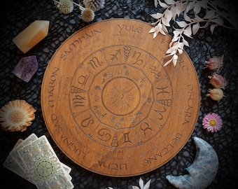 Könnte beinhalten: Ein Holzrad mit einem kreisförmigen Design, das die acht Sabbate des Radjahres zeigt. Das Rad ist mit den Namen der Sabbate beschriftet, darunter Yule, Imbolc, Ostara, Beltane, Litha, Lammas, Mabon und Samhain. Das Rad ist von verschiedenen Kristallen und anderen natürlichen Objekten umgeben.
