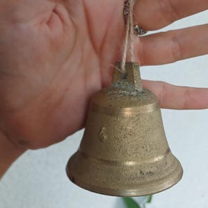 Op de afbeelding: Een kleine, verweerde koperen bel met een getextureerde, matte afwerking en een eenvoudig uitlopende rand. De bel heeft een kleine, gestempelde numerieke markering aan de zijkant en hangt aan een dunne, lichtgekleurde lus van natuurlijke vezels.