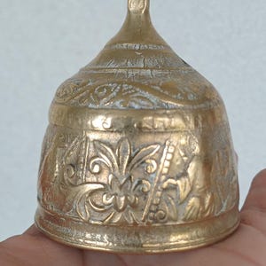 Op de afbeelding: Een goudkleurige koperen bel met een langwerpig handvat en een uitlopende voet. Het oppervlak van de bel is versierd met ingewikkelde reliëfpatronen van bloemen en gestileerde figuren, met een verweerde patina in de verdiepte delen.