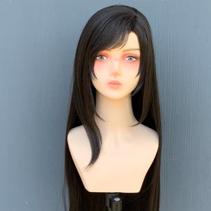 May include: A mannequin head with a long, black wig. The wig has a side part with bangs and long, straight hair. The mannequin has a pale complexion, with painted features including pink blush and blue eyes. The head is mounted on a black stand.
