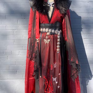 Könnte beinhalten: Ein rot-schwarzes, mehrlagiges Gewand mit schwarzem Kunstfellkragen, weiten Ärmeln und einem breiten schwarzen Gürtel mit silbernen rautenförmigen Akzenten. Das Kleidungsstück ist mit gestickten roten Flammen und weißen Schmetterlingsmotiven verziert.