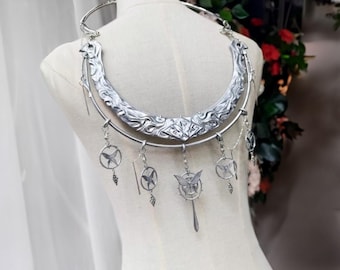 May include: Silver-toned statement necklace with intricate carved details and dangling charms. The necklace features a wide, curved collar with floral and geometric patterns. Several charms, including butterflies and circular designs, hang from the collar.