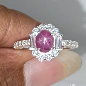 May include: A silver ring featuring a large oval, star-shaped ruby gemstone, surrounded by a halo of small, round, clear gemstones. The ring's band is also adorned with smaller, clear gemstones. The text "dkjewelrystore" is visible.