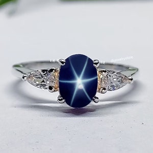 Pode incluir: Um anel de prata com uma gema de safira estrela azul e duas gemas transparentes menores. A safira estrela tem uma estrela de seis pontas no centro.