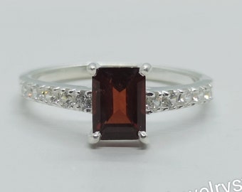 May include: A silver ring with a red gemstone and small clear stones around the band.