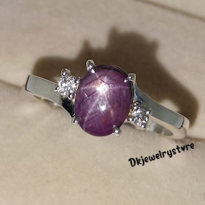 May include: A silver ring featuring a large, oval, star-shaped, purplish-red gemstone as the centerpiece. Two smaller, clear gemstones flank the main stone. The ring is set against a neutral background.