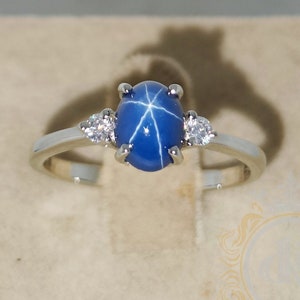 Pode incluir: Um anel de prata com uma gema de safira azul estrelada e duas gemas menores transparentes. O anel está colocado sobre um fundo branco.