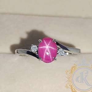 May include: A silver ring with a pink star sapphire and two smaller diamonds. The ring is set in a white gold band and is displayed in a red velvet box.