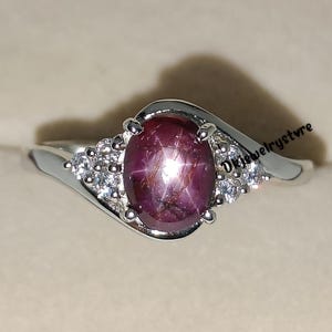 May include: A silver ring featuring a large, oval-cut, star ruby gemstone. The ruby is a deep red color with a star-like effect. The ring has a delicate, swirling design with small, clear accent stones.