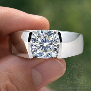 May include: A silver ring featuring a large, square-cut, clear gemstone. The ring has a wide band and a modern design. The gemstone is faceted, reflecting light. The ring is held in a hand against a blurred green background.