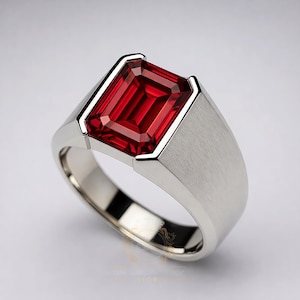 Könnte beinhalten: Ein silberner Ring mit einem großen, rechteckigen, tiefroten Edelstein. Der Ring hat eine gebürstete Metalloberfläche am Band und eine polierte Lünettenfassung um den Stein. Der Edelstein hat eine lebendige, auffällige Farbe.