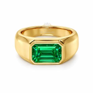 Può includere: Un anello d'oro con una gemma verde taglio smeraldo rettangolare. L'anello ha una fascia spessa e arrotondata e un'incastonatura a castone. L'anello è su sfondo bianco.