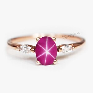 May include: Rose gold ring featuring a large, oval-cut, star ruby gemstone with a six-ray star pattern. Two smaller, marquise-cut, clear gemstones flank the ruby. The ring has a delicate band and is set against a white background.