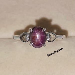 May include: A silver ring featuring a large, oval, star ruby gemstone. The ruby is a deep reddish-purple color with a star-like effect. Two smaller, heart-shaped gemstones flank the ruby. The ring is set against a neutral background.