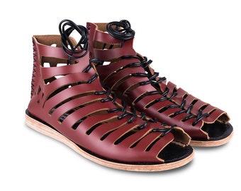 May include: A pair of burgundy leather sandals with open toes and a cage-like design. The sandals feature horizontal straps and vertical cutouts, laced with black cords. The soles are light-colored, and the sandals are set against a white background.