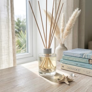 May include: A clear glass reed diffuser bottle with a light grey cap, containing liquid and small seashells at the base. Several wooden reeds extend upwards from the bottle. A small beige starfish sits nearby, next to a stack of four hardcover books.
