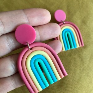 K&ouml;nnte beinhalten: Ein Paar Regenbogen-Ohrringe aus Polymer Clay. Jeder Ohrring hat einen Regenbogenbogen mit einem rosa Kreis oben. Der Regenbogenbogen besteht aus sechs verschiedenen Farben: Rot, Orange, Gelb, Gr&uuml;n, Blau und Lila.