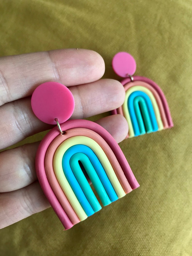 K&ouml;nnte beinhalten: Ein Paar Regenbogen-Ohrringe aus Polymer Clay. Jeder Ohrring hat einen Regenbogenbogen mit einem rosa Kreis oben. Der Regenbogenbogen besteht aus sechs verschiedenen Farben: Rot, Orange, Gelb, Gr&uuml;n, Blau und Lila.