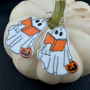 Könnte beinhalten: Ein Paar Halloween-Ohrringe. Jeder Ohrring zeigt ein weißes Gespenst mit schwarzen Augen, das ein orangefarbenes Buch und einen Kürbis hält. Die Ohrringe haben silberfarbene Fischhaken-Ohrdrähte. Die Ohrringe sind auf einem weißen Kürbis platziert.