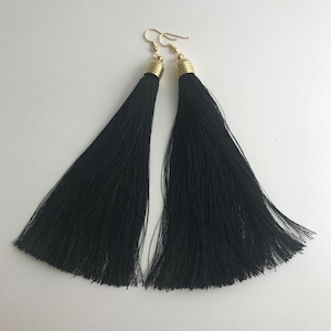 Könnte beinhalten: Ein Paar lange, schwarze Quastenohrringe. Die Quasten bestehen aus feinen, seidigen Fäden und sind an goldfarbenen Ohrhaken befestigt. Die Ohrringe haben ein schlichtes, elegantes Design.