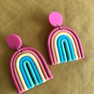 K&ouml;nnte beinhalten: Ein Paar regenbogenfarbene, bogenf&ouml;rmige Ohrringe mit rosa Kreis-Akzenten. Die Ohrringe sind aus Polymer Clay gefertigt und haben eine matte Oberfl&auml;che.