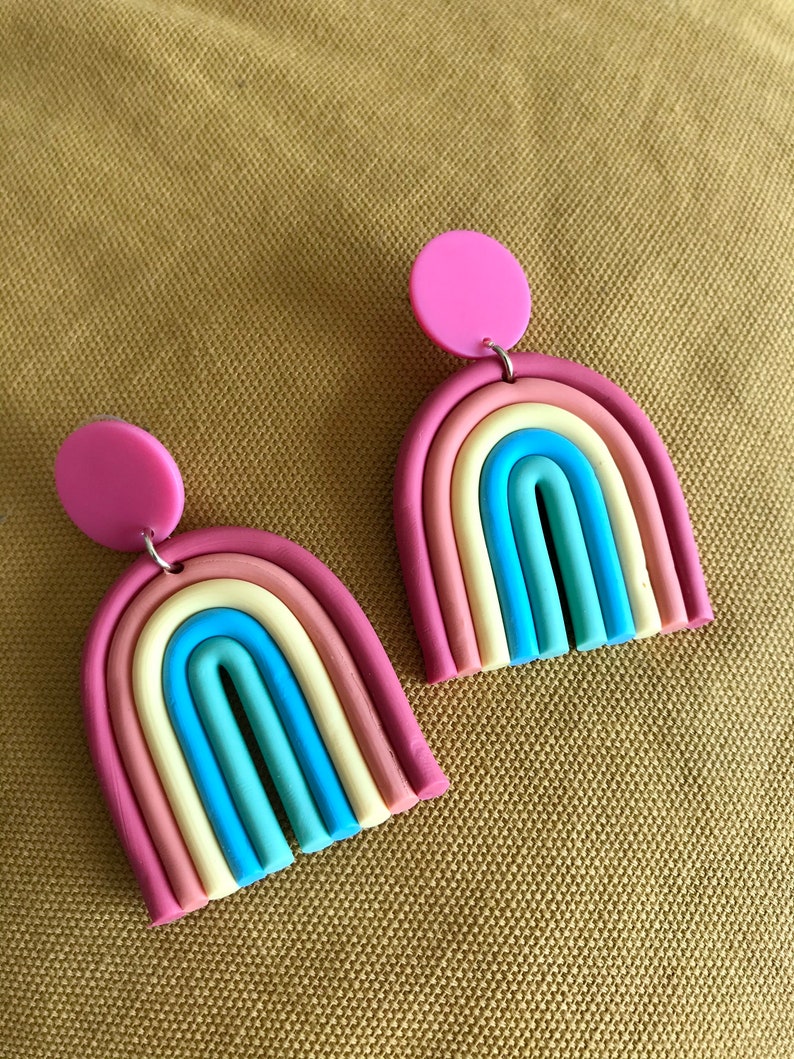 K&ouml;nnte beinhalten: Ein Paar regenbogenfarbene, bogenf&ouml;rmige Ohrringe mit rosa Kreis-Akzenten. Die Ohrringe sind aus Polymer Clay gefertigt und haben eine matte Oberfl&auml;che.