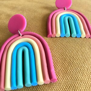 K&ouml;nnte beinhalten: Ein Paar regenbogenfarbene Ohrringe mit rosa Oberteilen. Die Ohrringe sind aus Polymer Clay gefertigt und haben die Form von Regenb&ouml;gen. Jeder Ohrring hat sechs Farbschichten: rosa, pfirsich, creme, hellblau, blau und gr&uuml;n.