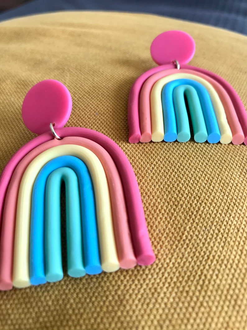 K&ouml;nnte beinhalten: Ein Paar regenbogenfarbene Ohrringe mit rosa Oberteilen. Die Ohrringe sind aus Polymer Clay gefertigt und haben die Form von Regenb&ouml;gen. Jeder Ohrring hat sechs Farbschichten: rosa, pfirsich, creme, hellblau, blau und gr&uuml;n.