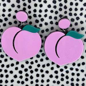 Könnte beinhalten: Ein Paar rosa Pfirsich-Ohrringe mit grünen Blättern. Die Ohrringe befinden sich auf einem schwarz-weißen Polka-Dot-Hintergrund.