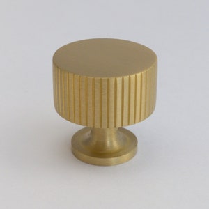 May include: A gold-colored cabinet knob with a cylindrical body and a ridged texture. The knob has a small, circular base and a flat top. This hardware is suitable for drawers and cabinets.